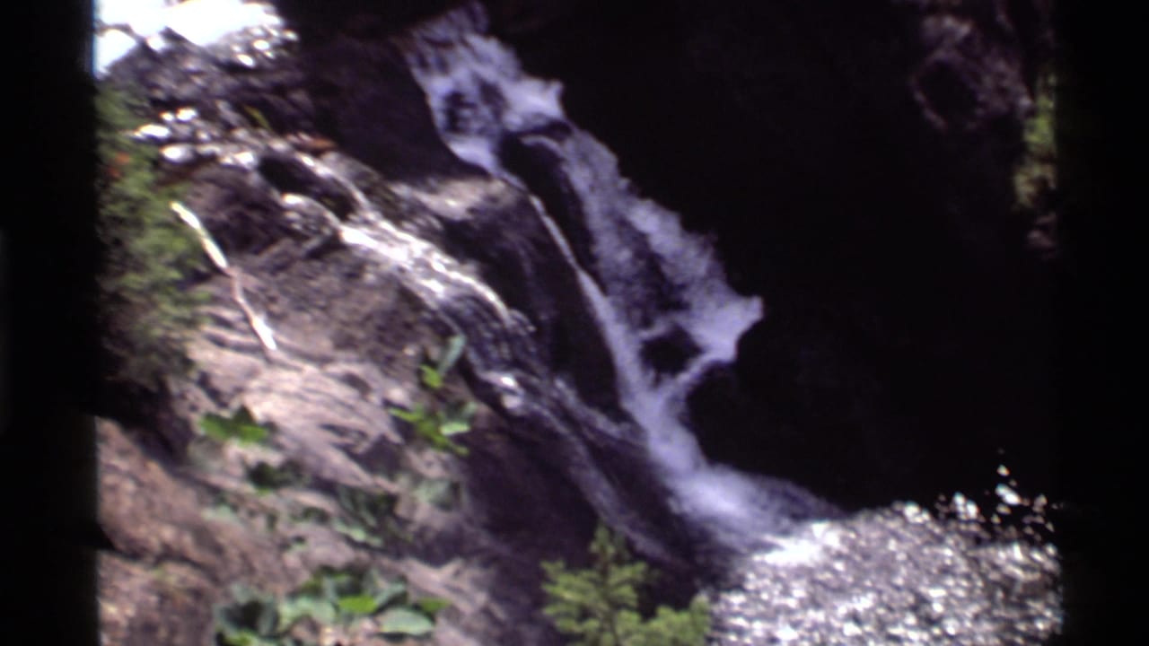 Waterfall spilling over dark rock into sunlit water with green plants nearby.