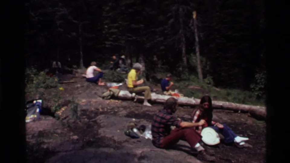 Adults relax in a forest clearing at Holland Lake, Montana, in 1972, sitting on rocks and a fallen log with bags and food nearby.