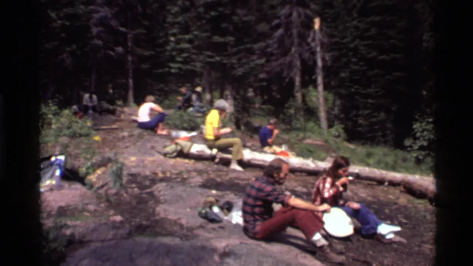 Several adults sit and talk in a sunlit pine forest near Holland Lake, Montana, with 1970s clothing and picnic items visible.