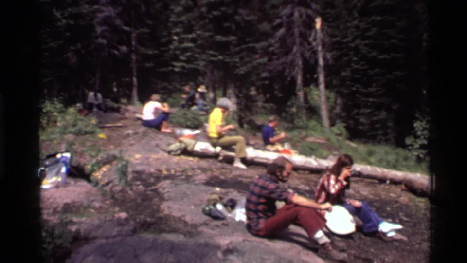 Adults in casual 1970s clothing sit near a fallen log in a Holland Lake forest clearing, with bags and picnic items on the ground.