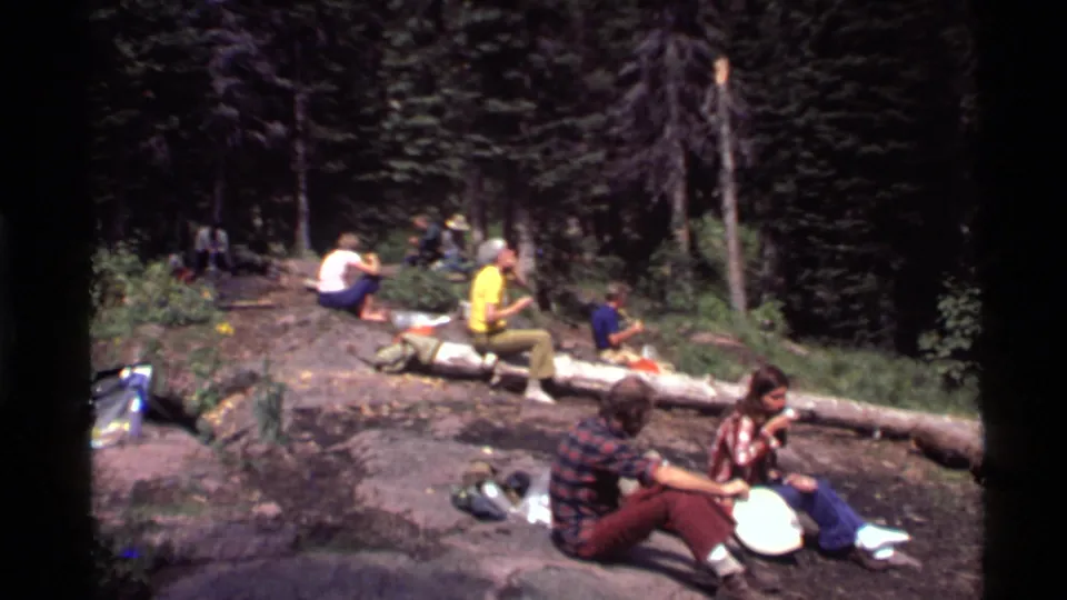 The group continues relaxing in the Holland Lake forest, seated on rocks and beside a log with food and gear spread nearby.