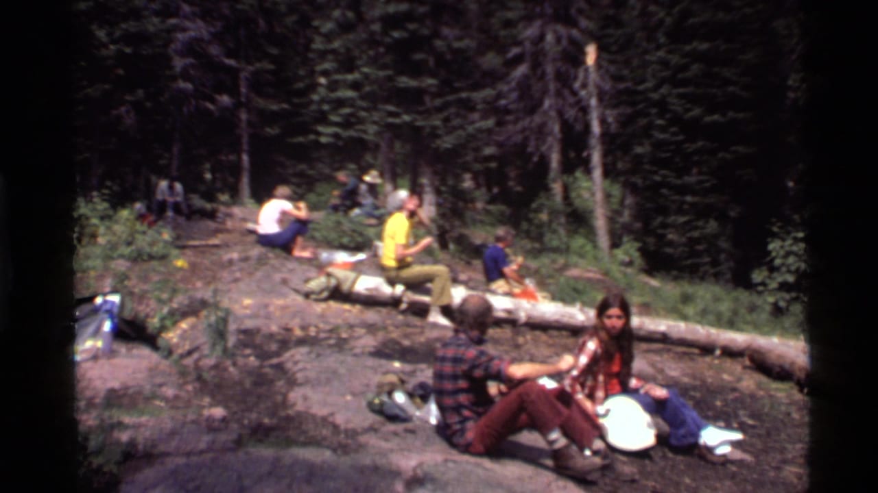 Young adults in 1970s clothing sit on logs and rocks during a camping stop near Holland Lake, Montana.