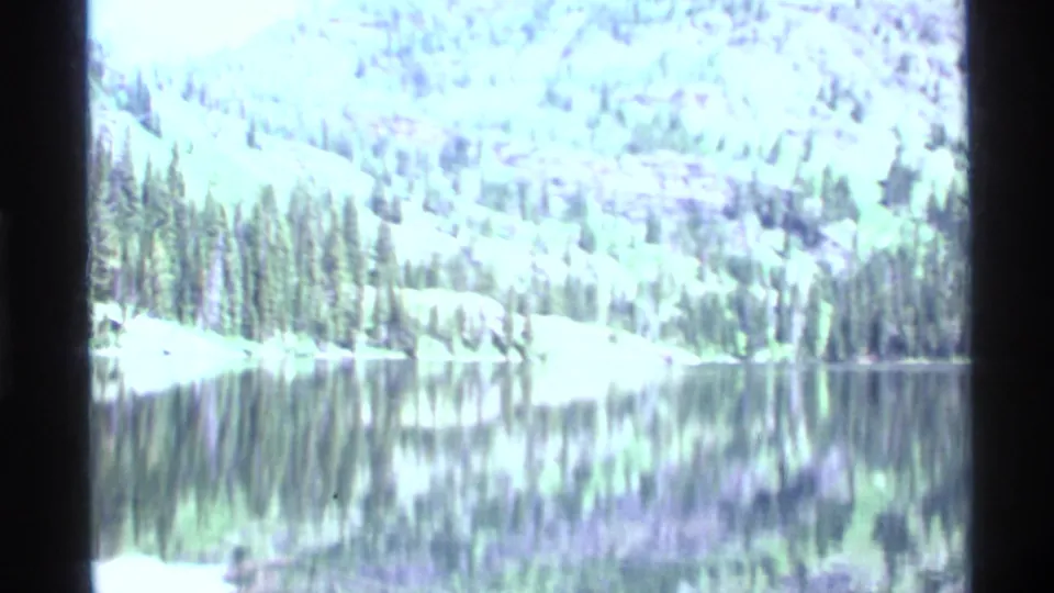 Holland Lake appears calm, reflecting evergreen trees and a wooded mountain slope in 1972 home-movie footage.