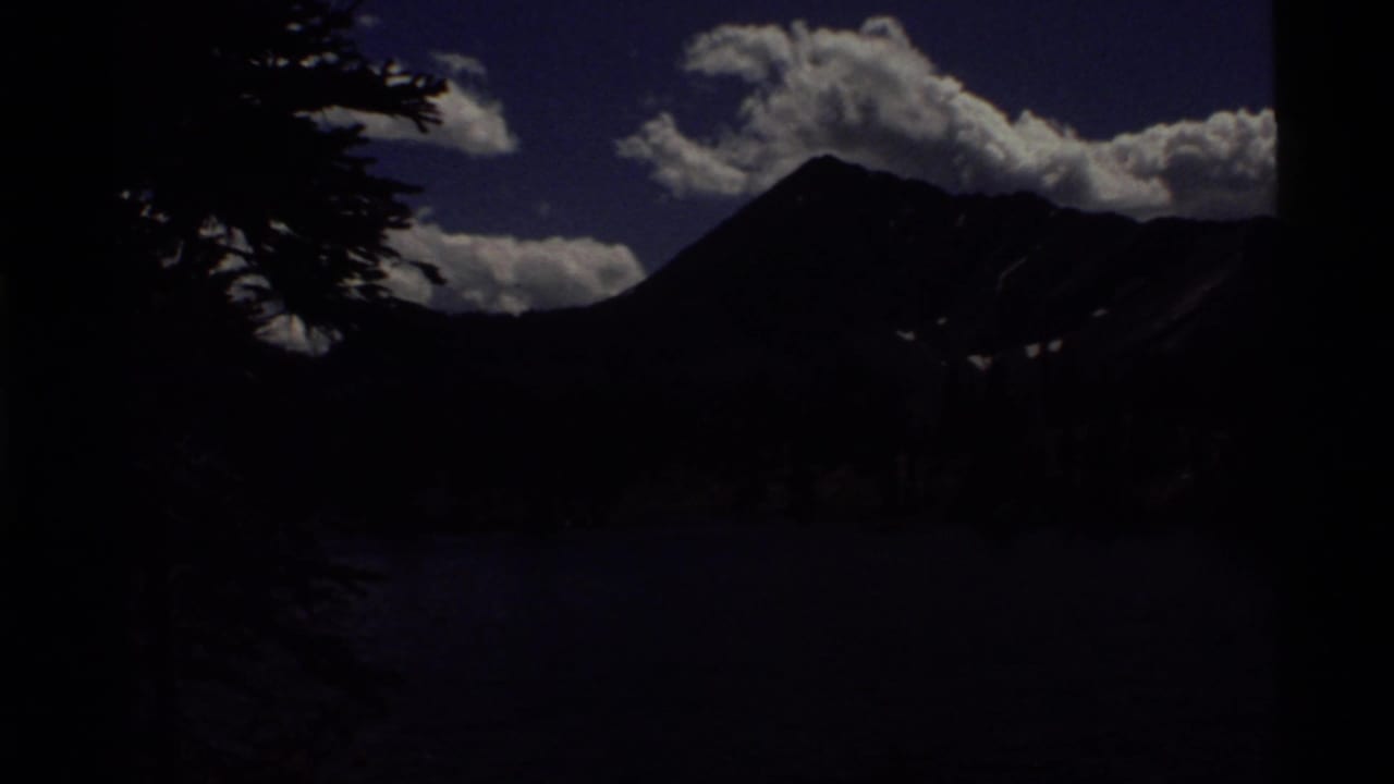 Underexposed 1972 8mm view across Holland Lake, Montana, with dark tree silhouettes, mountain peaks, clouds, and blue sky.
