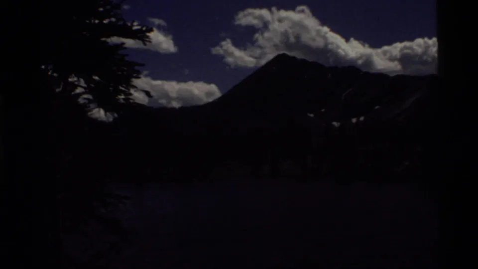 Underexposed 1972 8mm view across Holland Lake, Montana, with dark tree silhouettes, mountain peaks, clouds, and blue sky.
