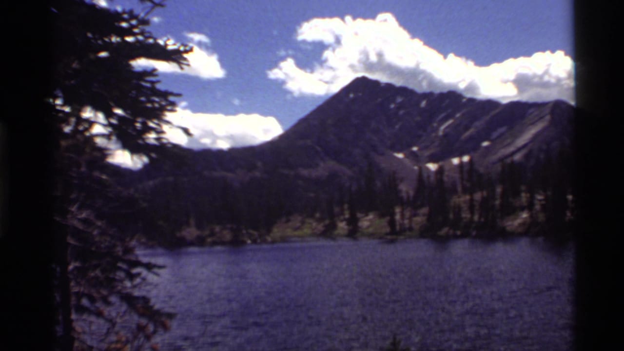 Mountain lake with evergreen shore, rocky peaks, white clouds, and blue sky.