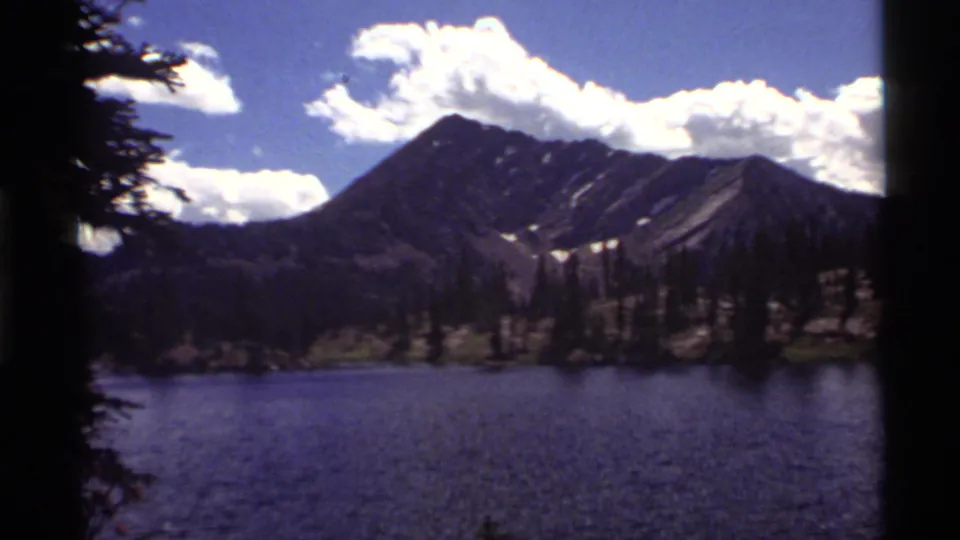 Wide 1972 home-movie frame of Holland Lake, Montana, with blue water, evergreen trees, mountain slopes, snow patches, and bright clouds.