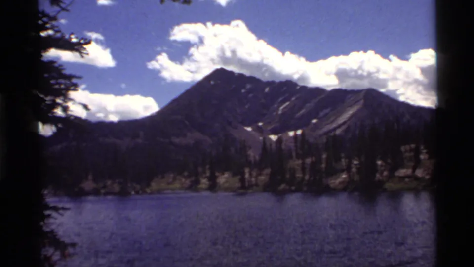 1972 8mm Holland Lake, Montana, frame with dark tree edges, rippled lake water, evergreen shore, rugged peaks, and sunlit clouds.
