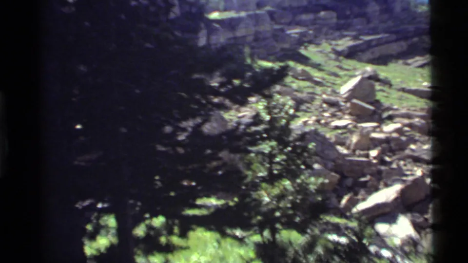 1972 Holland Lake frame with darker exposure, evergreen foliage, green ground cover, rocks, and debris on a rugged slope.