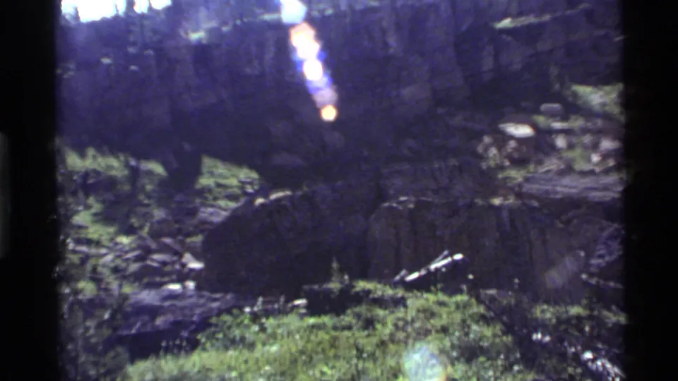 Darker 1972 8mm frame at Holland Lake with grass, rocky ground, fallen wood, and vertical sunlight flare.