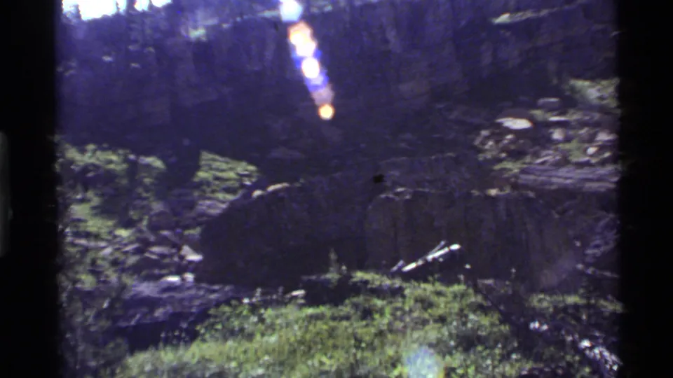 1972 Holland Lake 8mm frame showing a shadowed rocky grass slope, fallen wood, and bright flare across the upper frame.