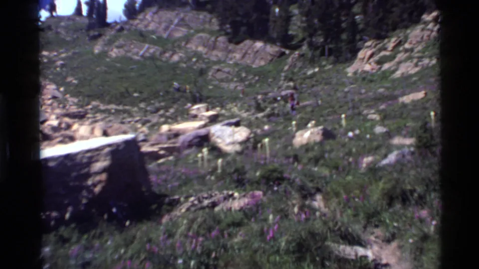 Wide 1972 8mm view of a rocky, vegetated slope near Holland Lake with distant hikers moving through the landscape.