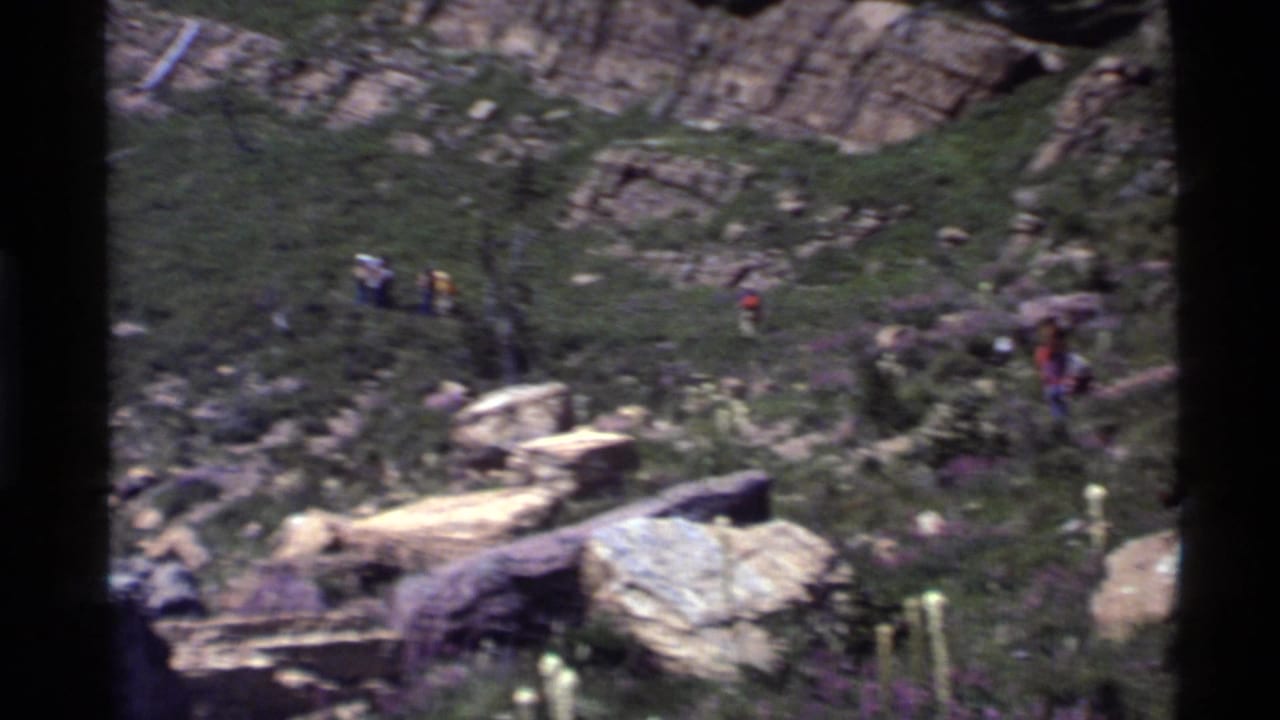 Small hikers on a rocky green mountain slope below canyon walls.