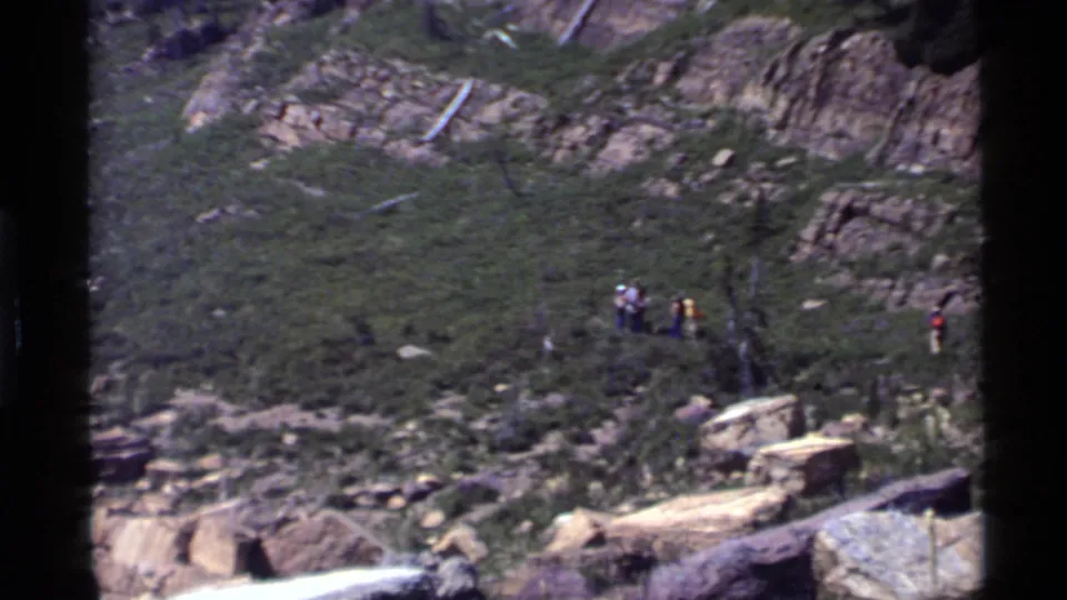 Home-movie frame of hikers spread across a rugged Holland Lake mountain slope, with rocks, brush, and canyon wall visible.
