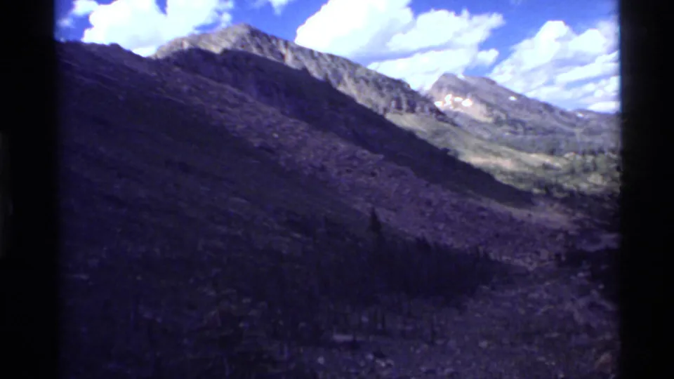 Wide 1972 8mm mountain view near Holland Lake, Montana, with shadowed valley slopes, distant peaks, blue sky, and white clouds.