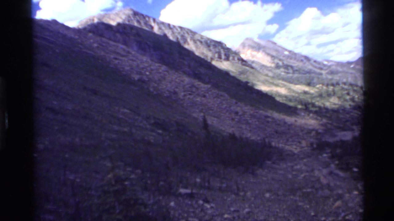 Mountain valley with rocky slopes, conifer trees, and a bright clouded sky.