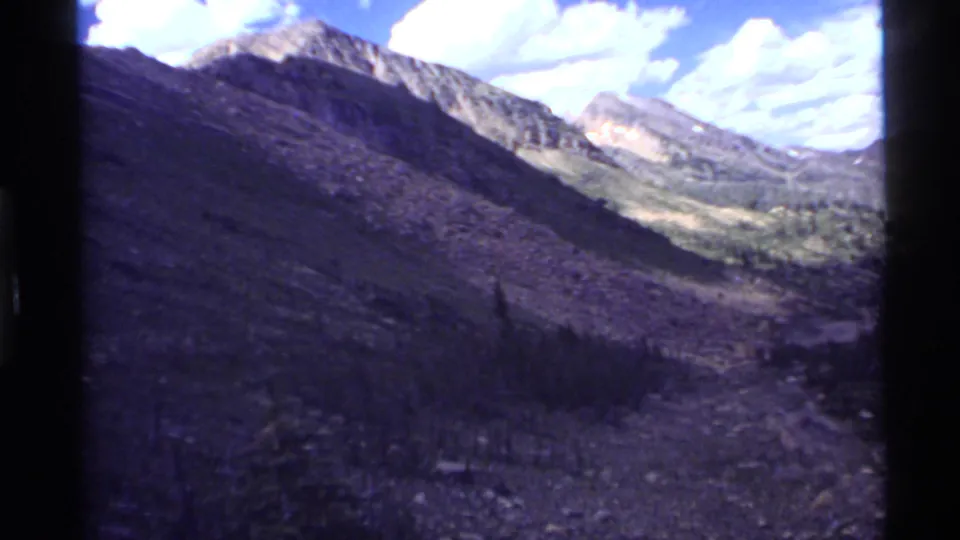 1972 8mm home-movie view from Holland Lake, Montana, showing rugged mountain slopes, a shadowed valley, and scattered clouds.