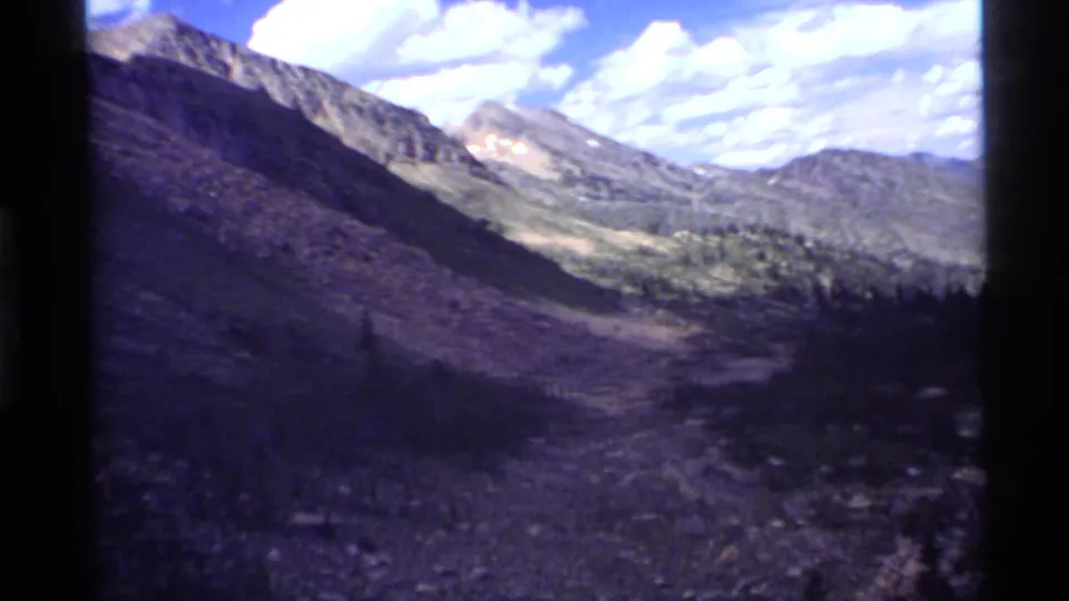 1972 Holland Lake, Montana footage shows a sunlit ridge, rocky terrain, conifer trees, and a bright sky within 8mm frame edges.