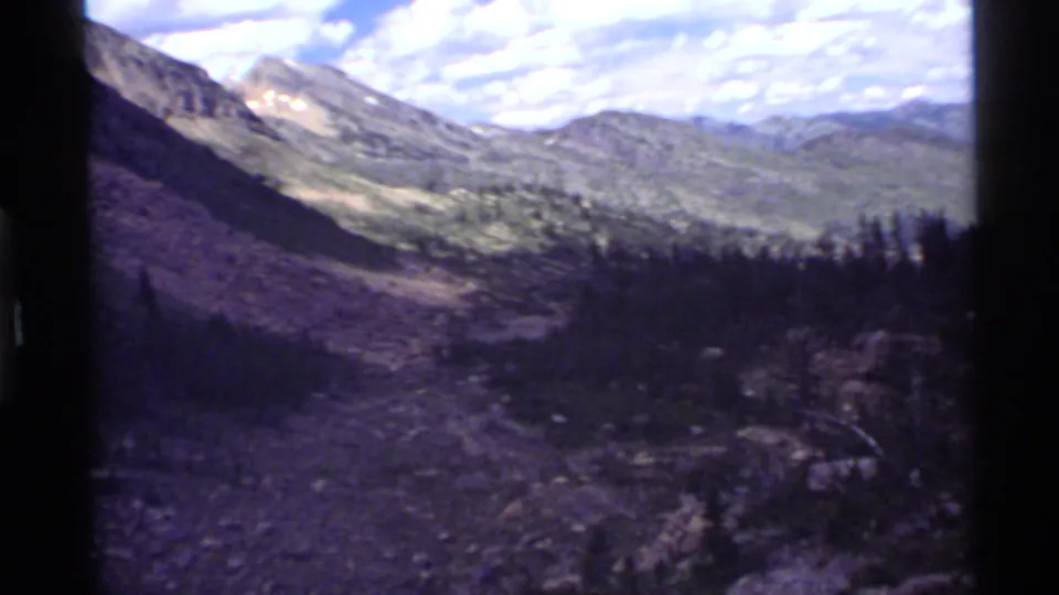 1972 8mm view near Holland Lake, Montana, with rocky valley terrain, green scrub, conifers, and distant mountain ridges.