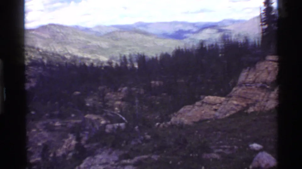 Handheld 8mm frame of the Holland Lake area with mountain ridges, dense conifers, and rocky foreground terrain.