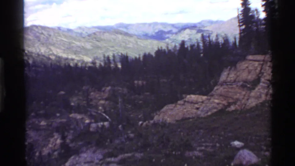 1970s home-movie frame near Holland Lake showing rocky terrain, trees, and layered mountain slopes in soft 8mm color.