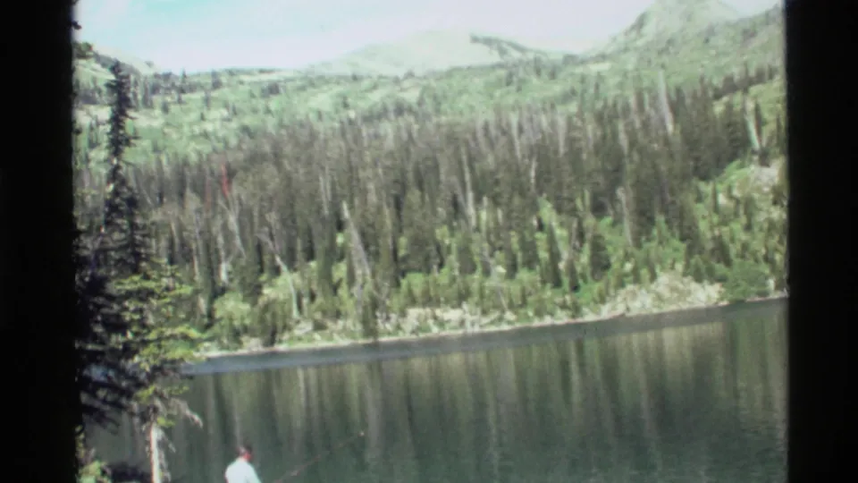 1972 8mm frame near Holland Lake, Montana, with a fisherman at the shore, calm water, conifers, and mountain slopes.