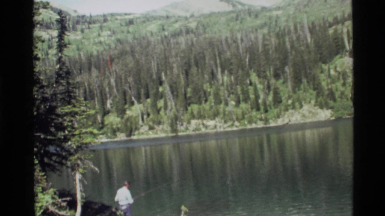 A man stands low in frame near Holland Lake, Montana, with calm water, evergreen forest, and mountain slopes visible in 1972 8mm footage.