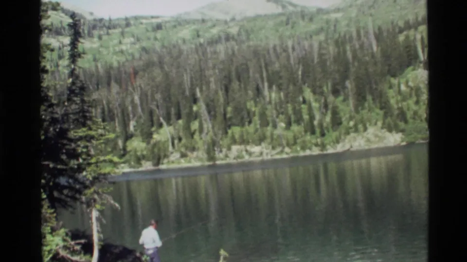 A man stands low in frame near Holland Lake, Montana, with calm water, evergreen forest, and mountain slopes visible in 1972 8mm footage.
