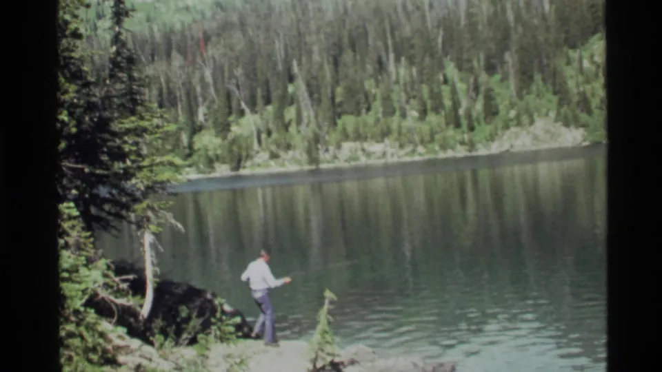 The fisherman stands at the Holland Lake shoreline, holding a rod toward the water with trees and reflected slopes across the lake.