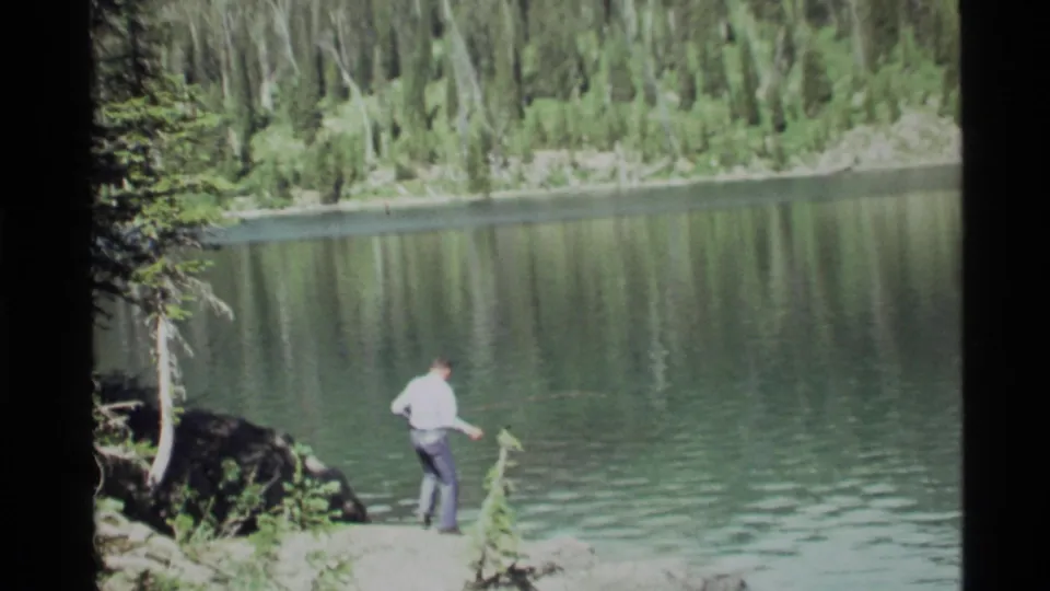 A man in a light shirt and dark pants stands near the waterline at Holland Lake, framed by rocks, trees, and rippling water.