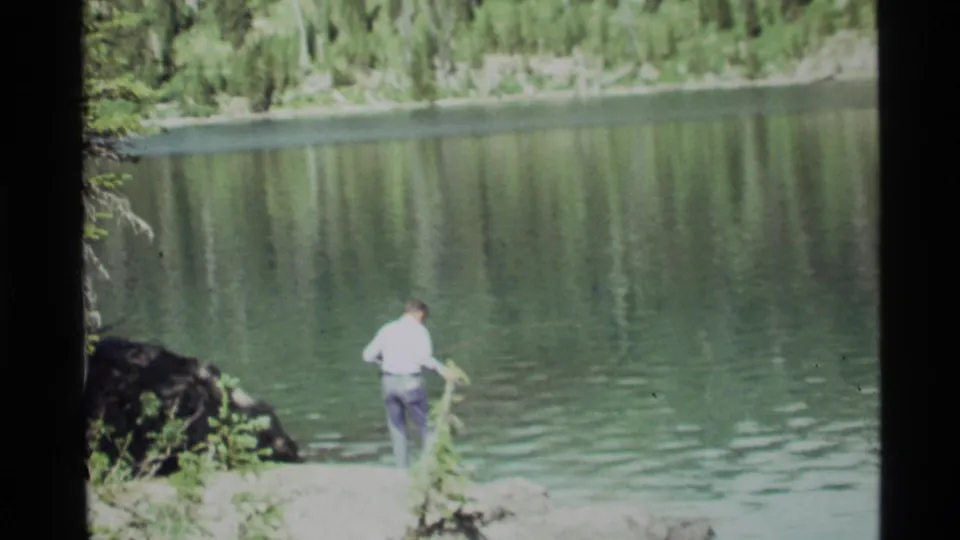 The fisherman faces Holland Lake from the rocky edge as the 8mm frame catches water ripples, reflected trees, and a wooded opposite shore.