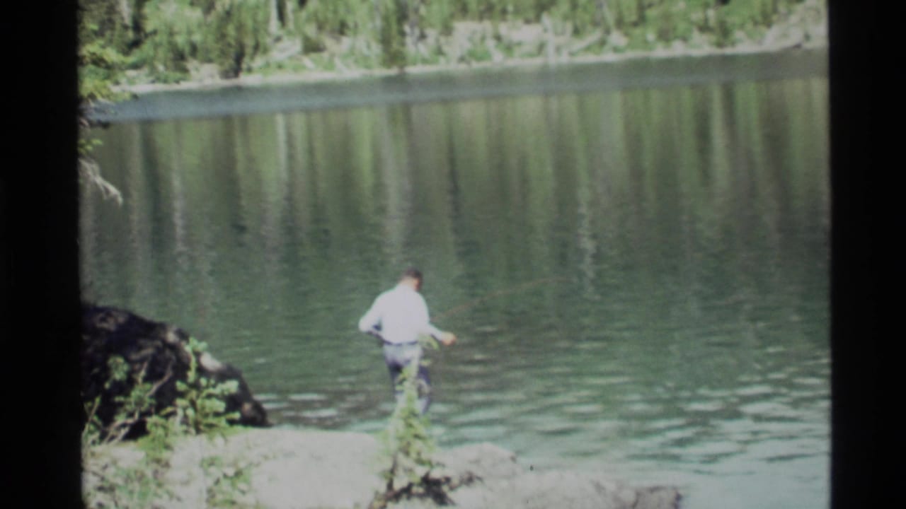 At Holland Lake, Montana in 1972, a man stands on a rocky shore and casts a fishing rod toward the still water.