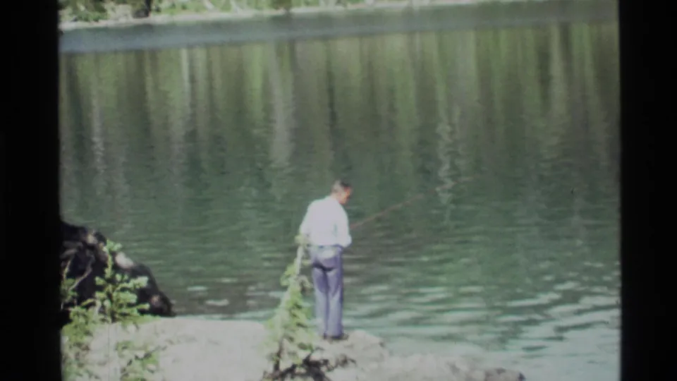 The fisherman faces Holland Lake with his rod angled across the water, framed by rocks, greenery, and reflected trees.