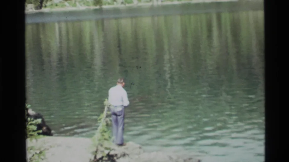 A rear view shows the man standing still at Holland Lake, fishing beside rocks and leafy plants with calm water ahead.