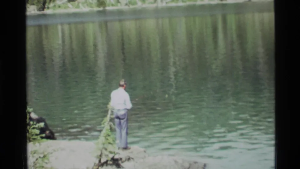 The 1972 home-movie frame shows the fisherman paused at the water's edge, with Holland Lake and tree reflections filling the background.
