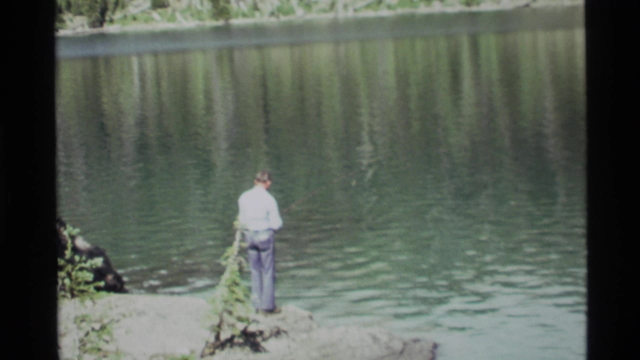 Man stands on a rocky lakeshore holding a fishing pole over calm water.