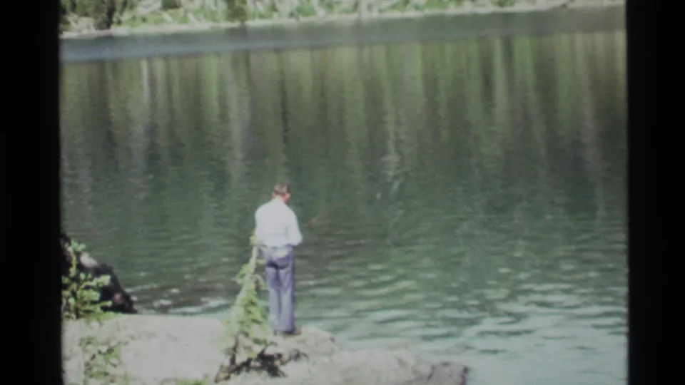 At Holland Lake, Montana in 1972, a man in a light shirt and dark pants stands on rocks while fishing from the shore.