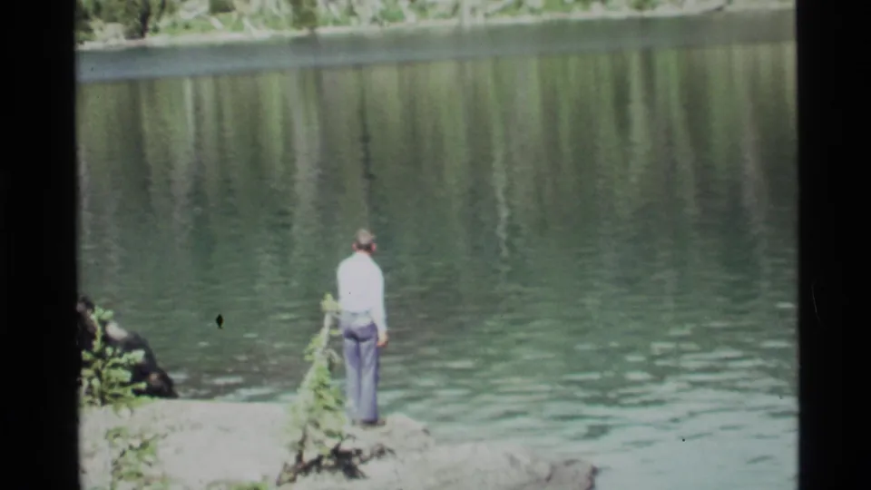 A 1972 8mm frame shows a man at Holland Lake facing the water with a fishing line cast from a rocky bank.