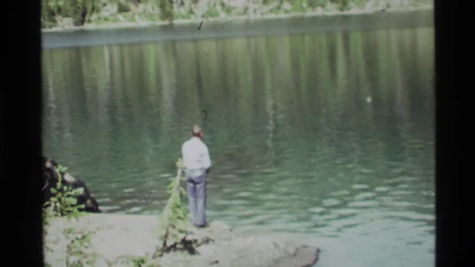 The man remains on the rocky Holland Lake shoreline, fishing beside low greenery and softly rippling water in this 1972 home-movie frame.