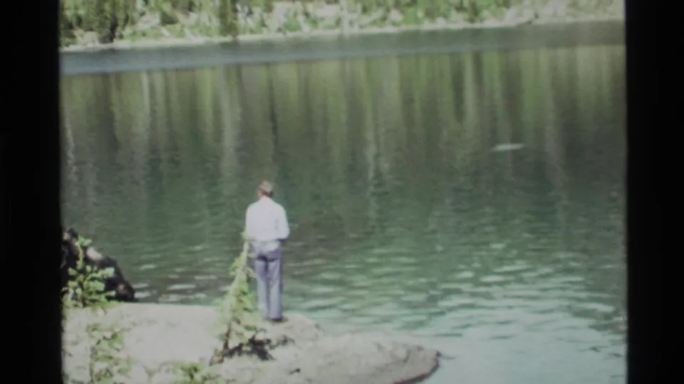 A man in a light shirt and dark trousers fishes from the edge of Holland Lake, Montana, with calm water filling most of the 1972 frame.