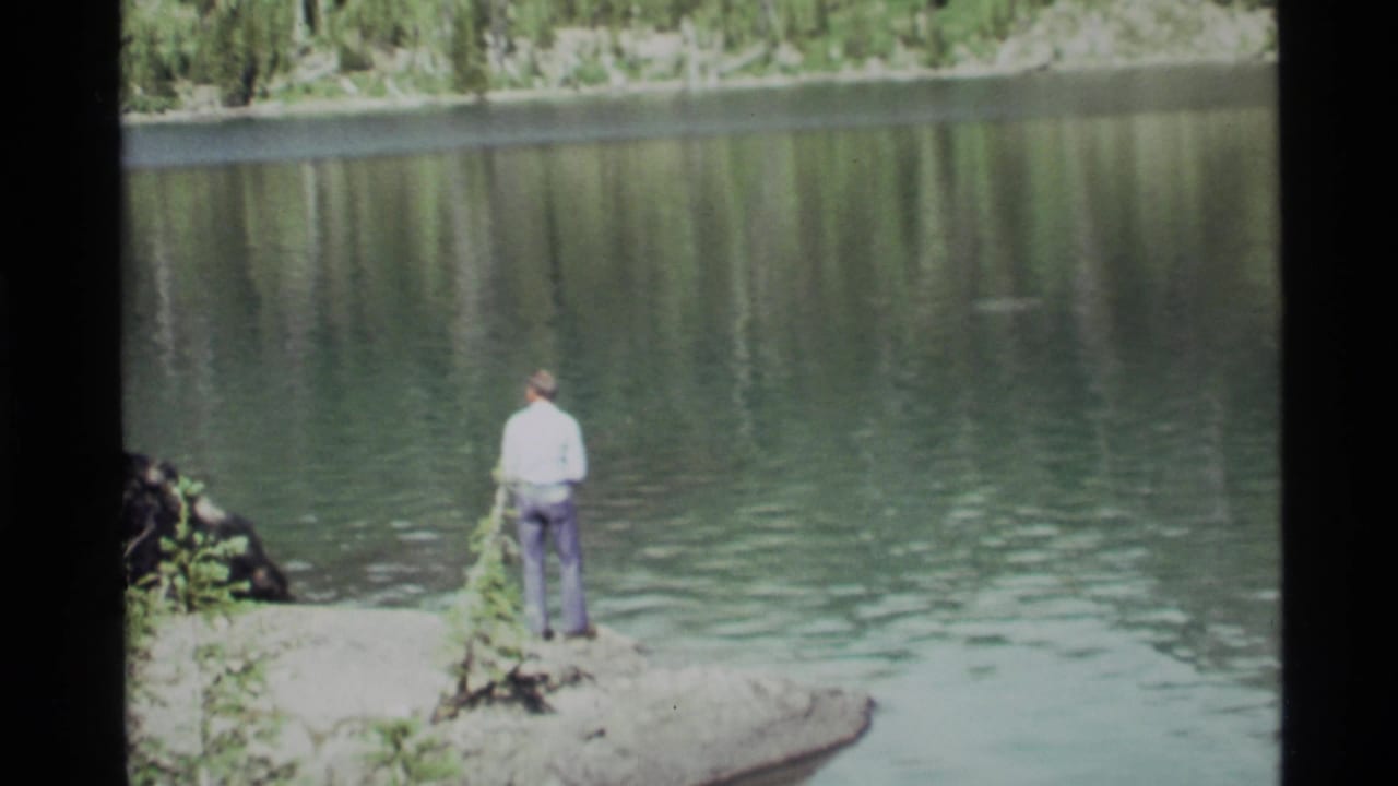 Man standing on a rocky lakeshore facing calm water and forest reflections.