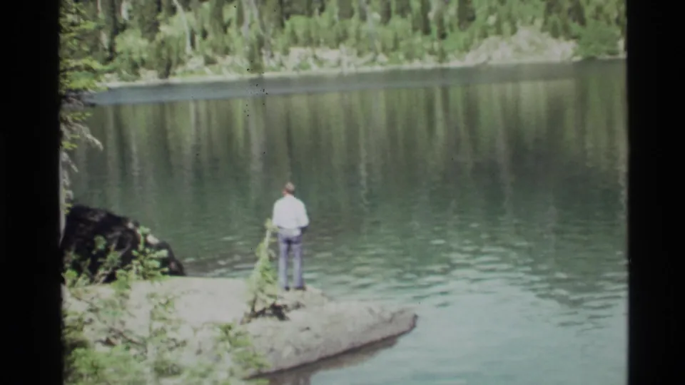 The 1972 Holland Lake view shows a man from behind on a small rocky point, with evergreen forest reflected in the water.