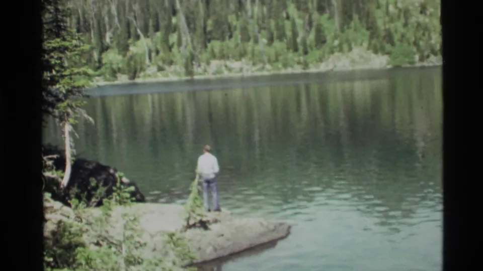 A lone adult stands near the waterline at Holland Lake, Montana, with rippled water and dense trees filling the frame.