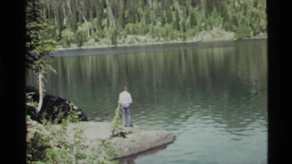 The final frame holds on the man at Holland Lake's rocky edge, facing the reflective water and forested opposite shore.