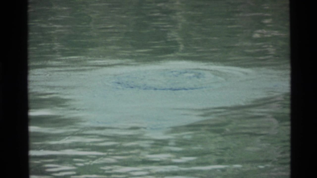 Holland Lake water in 1972 home-movie footage, showing a pale disturbance on the surface and dark scan borders.