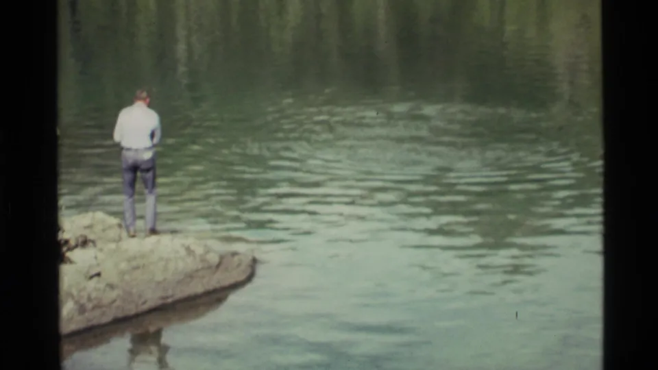 Adult man at Holland Lake, Montana, standing on a rock and facing green water in 1972 8mm footage.