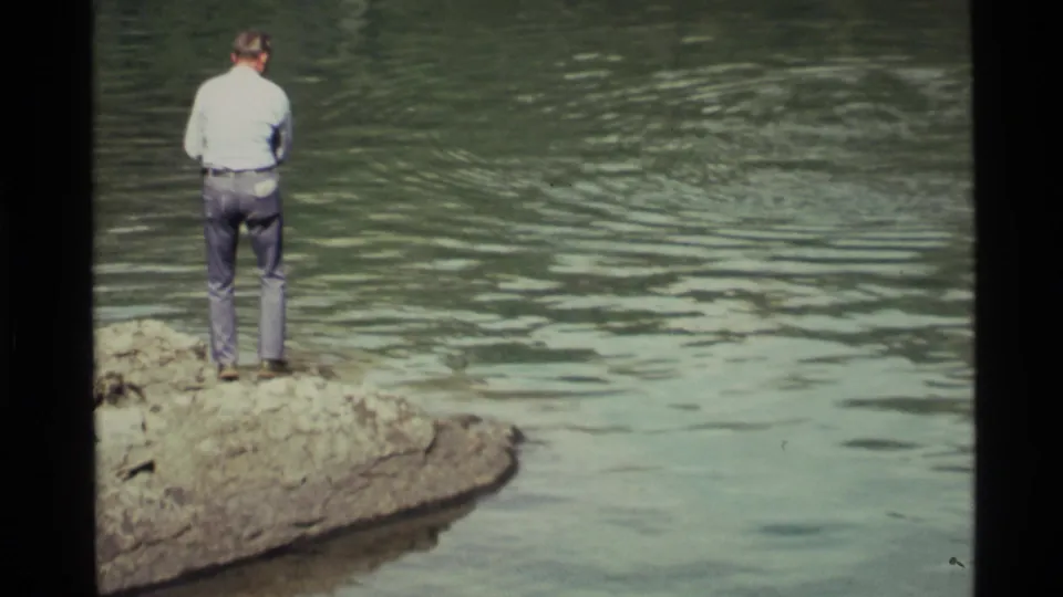 Adult man in a light shirt and jeans stands at the rocky shore of Holland Lake in 1972 home-movie footage.