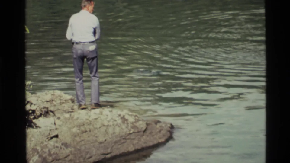 Closer 1972 Holland Lake frame showing a man from behind on a rocky edge, facing the rippled water.