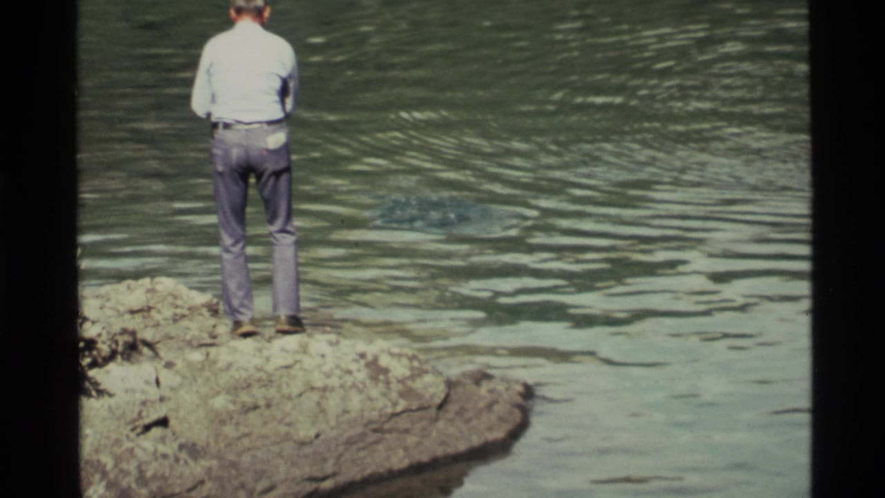 At Holland Lake, Montana in 1972, a man seen from behind stands on a rocky shore while ripples spread across the green-blue water.