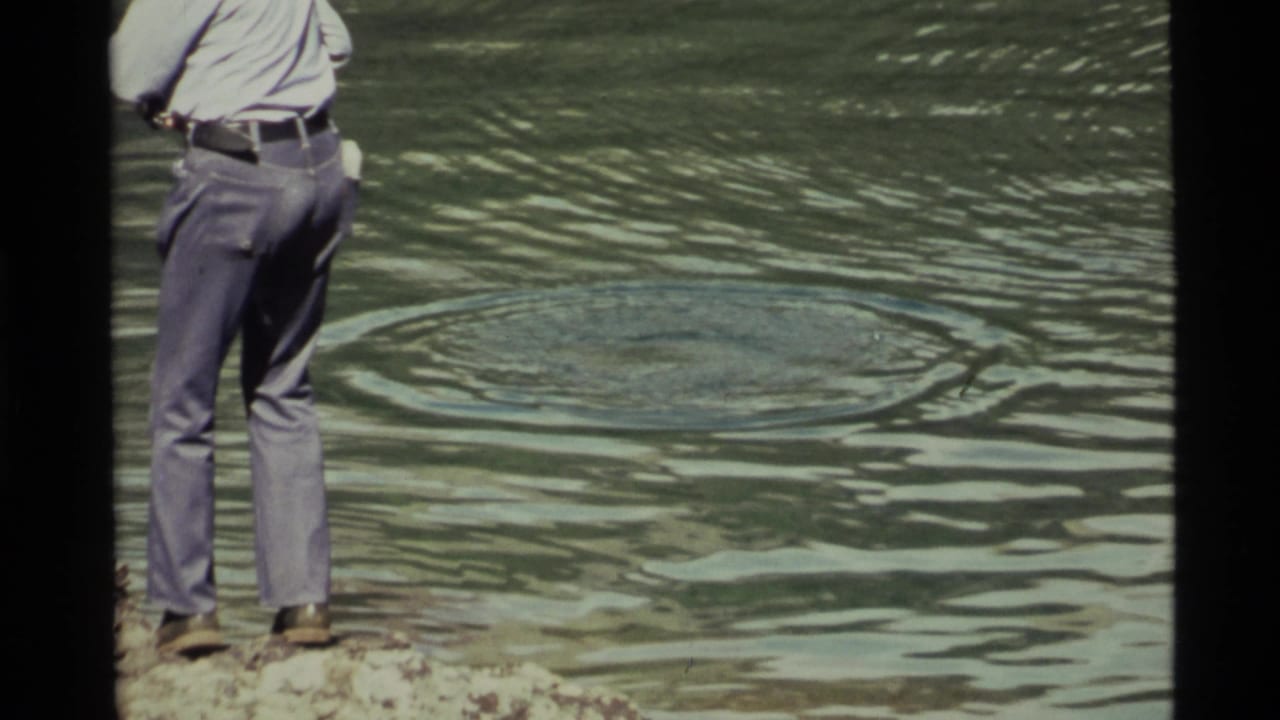 Closer rear view of a man on rocks facing circular ripples in the lake.